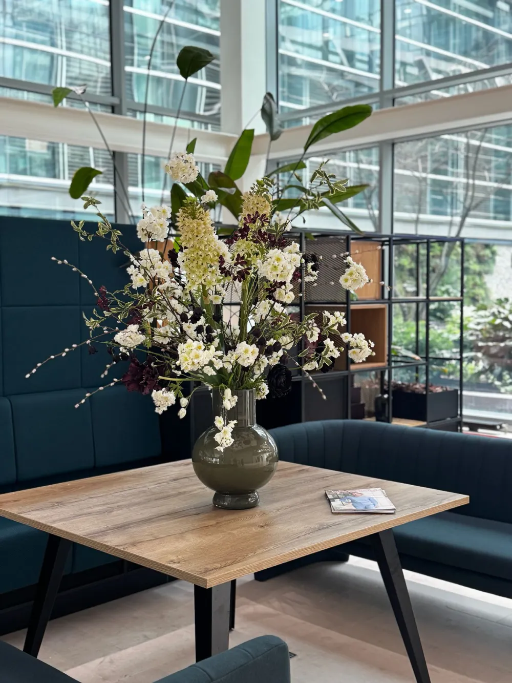Hoog zijden bloemstuk in een glanzende olijfgroene vaas op een houten vergadertafel